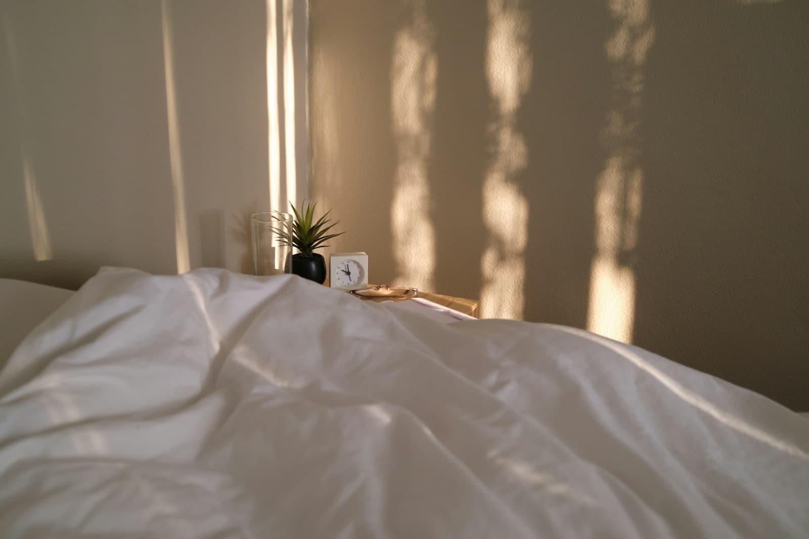 Morning light and window shadows across white bedding, with a small plant and a clock on the table beside it